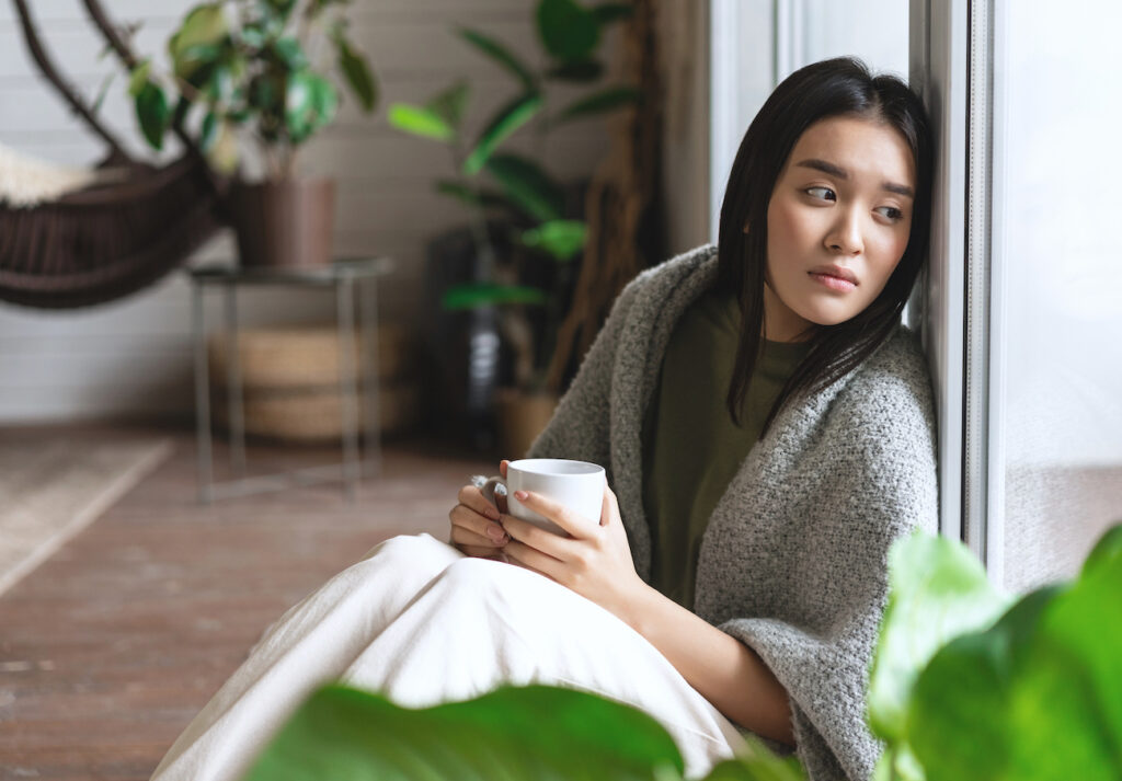 sad asian woman looking outside window sitting floor with miserable upset face drinking coffee